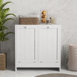 A white, double tilt-out laundry hamper with vertical panel doors and round knobs, topped with a wicker basket and decorative items. It stands beside a wire basket and a potted plant on a tiled floor.