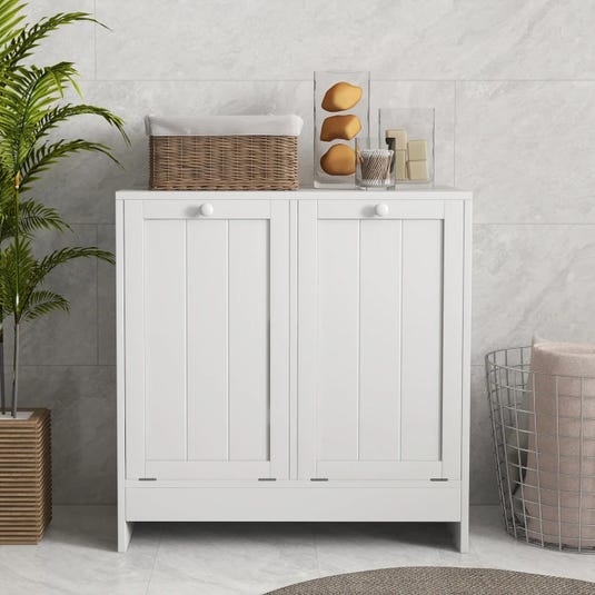 A white, double tilt-out laundry hamper with vertical panel doors and round knobs, topped with a wicker basket and decorative items. It stands beside a wire basket and a potted plant on a tiled floor.