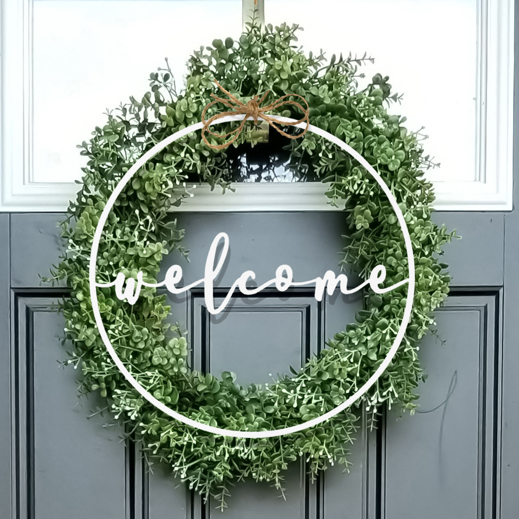 A door wreath with lush green foliage features a circular design. The word \“welcome\“ is elegantly written in white script across the center, with a small decorative bow at the top.