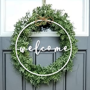 A door wreath with lush green foliage features a circular design. The word \“welcome\“ is elegantly written in white script across the center, with a small decorative bow at the top.