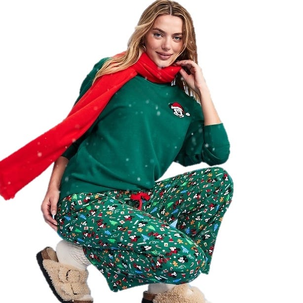A person wears a green sweater with a Santa design, green patterned pajama pants, a red scarf, and fluffy slippers, creating a festive and cozy outfit.