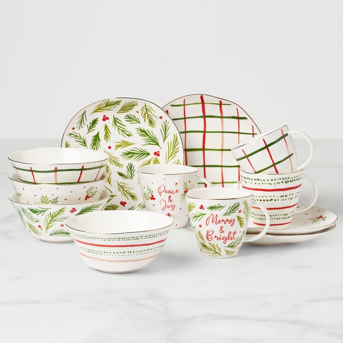 A holiday-themed dinnerware set featuring plates, bowls, and mugs with festive designs such as pine branches, berries, grid patterns, and phrases like \“Peace & Joy\“ and \“Merry & Bright\“ in red and green.