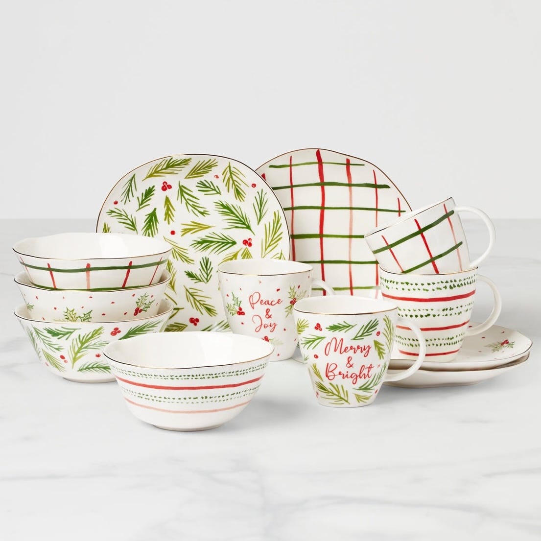 A holiday-themed dinnerware set featuring plates, bowls, and mugs with festive designs such as pine branches, berries, grid patterns, and phrases like \“Peace & Joy\“ and \“Merry & Bright\“ in red and green.