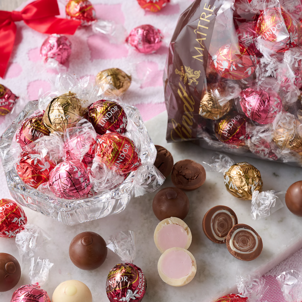 A variety of Lindt Lindor chocolate truffles are displayed, including red, pink, and gold-wrapped chocolates, some of which are cut open to show their creamy centers.