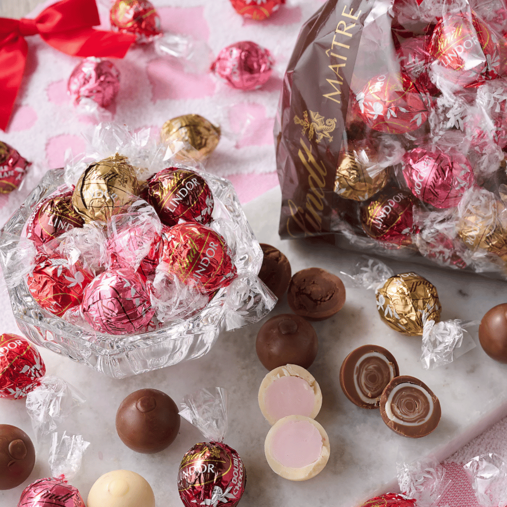 A variety of Lindt Lindor chocolate truffles are displayed, including red, pink, and gold-wrapped chocolates, some of which are cut open to show their creamy centers.