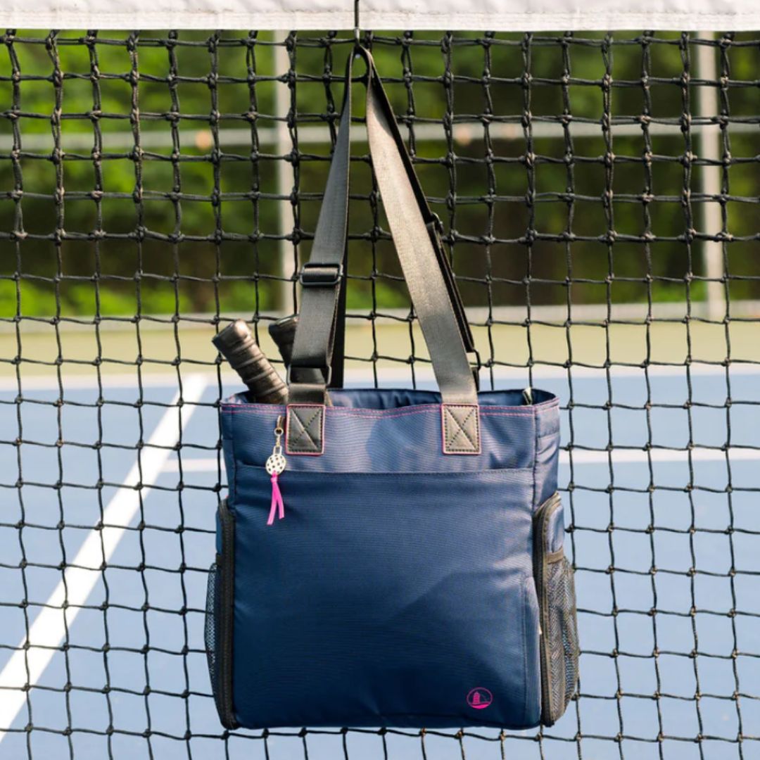 A navy tote bag with black straps is hanging on a tennis net, containing a tennis racket. It features side pockets and a small decorative charm.