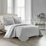 A gray, textured bedding set with a quilt and matching pillow shams in a neatly arranged, classic bedroom featuring an upholstered headboard, side table, and elegant decorative elements.