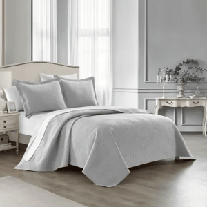 A gray, textured bedding set with a quilt and matching pillow shams in a neatly arranged, classic bedroom featuring an upholstered headboard, side table, and elegant decorative elements.