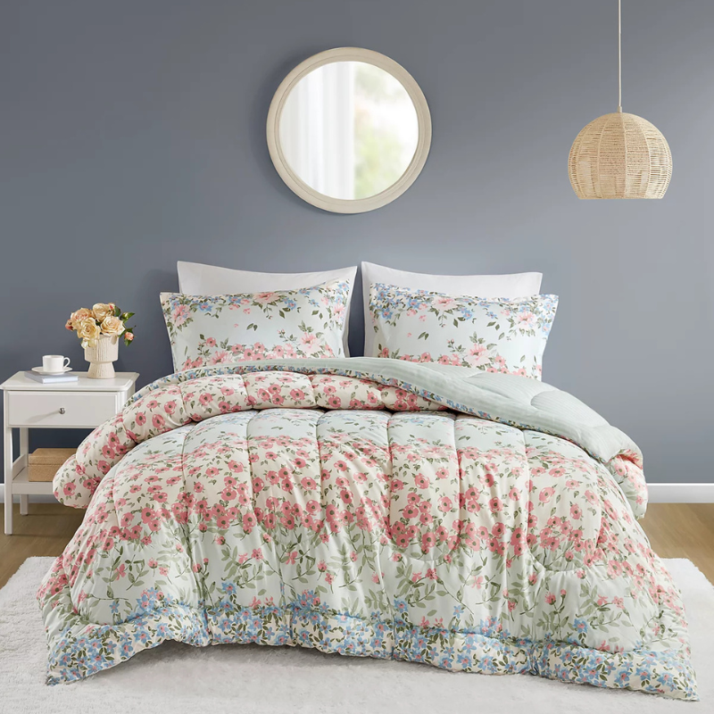 A bed is dressed with a floral-patterned comforter and matching pillows in a room featuring a round mirror and a wicker pendant light. A small white nightstand holds a vase of flowers and a cup.
