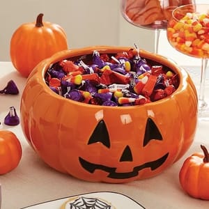 A pumpkin-shaped bowl filled with Halloween candy, including wrapped chocolates and candy corn. Additional pumpkins and a glass of candy corn are nearby on the table.