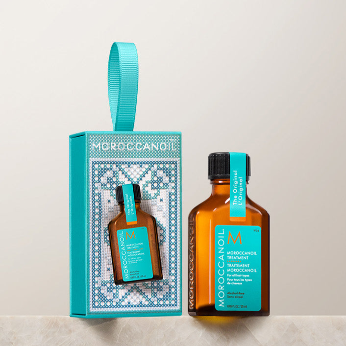 A small bottle of Moroccanoil Treatment and its packaging, featuring a teal and brown color scheme with a decorative design, is displayed on a marble surface.