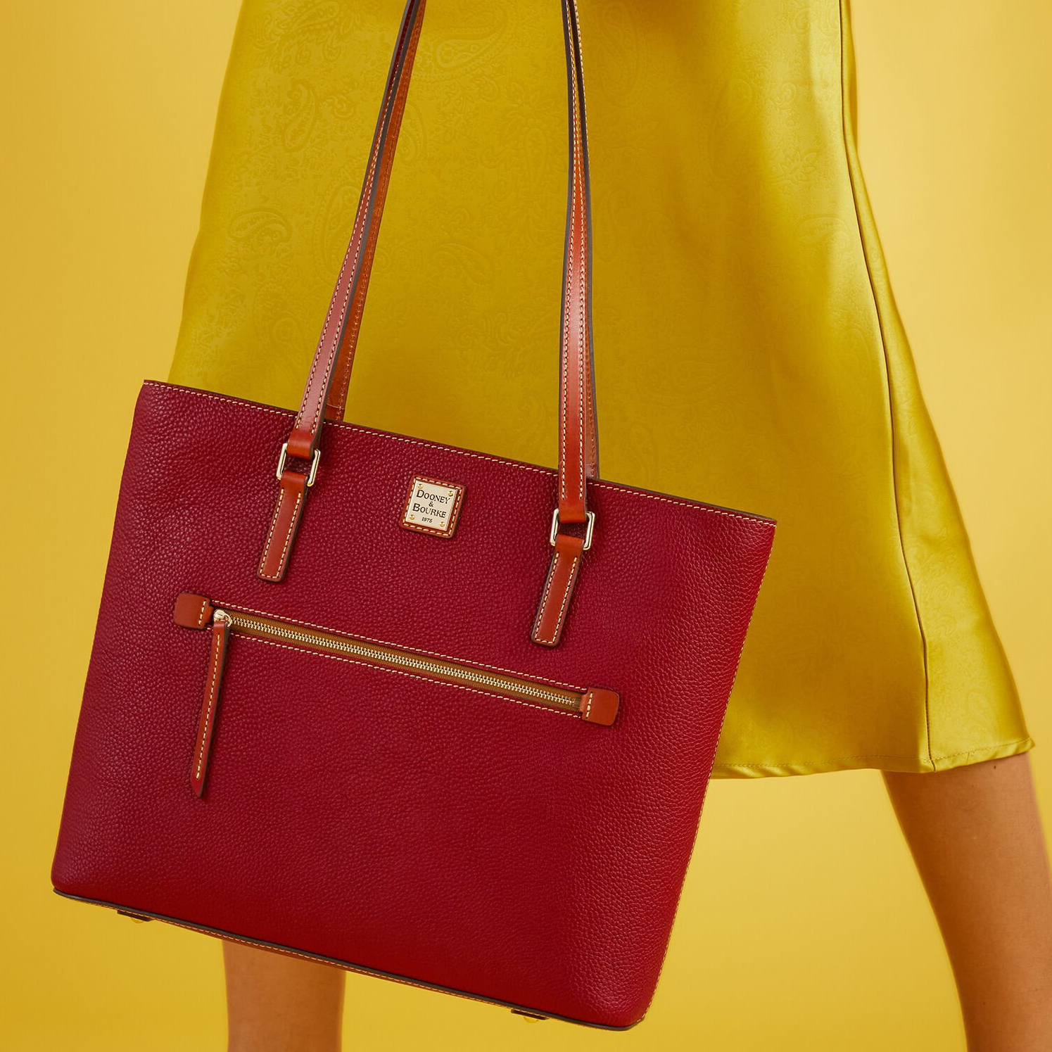 A red Dooney & Bourke tote bag with brown straps and a front zipper pocket, paired with a yellow background and a yellow skirt.