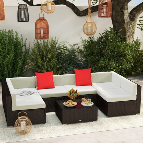 A modern outdoor patio set with a U-shaped sectional sofa featuring white cushions and two red throw pillows, accompanied by a dark rattan coffee table. Pendant and standing lanterns add decorative touches, while a tree and shrubs provide a natural backdrop.