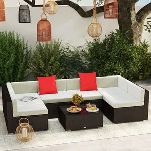 A modern outdoor patio set with a U-shaped sectional sofa featuring white cushions and two red throw pillows, accompanied by a dark rattan coffee table. Pendant and standing lanterns add decorative touches, while a tree and shrubs provide a natural backdrop.
