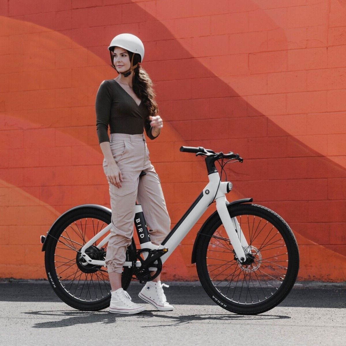 A woman stands beside an electric bicycle against an orange wall, wearing casual clothing and a white helmet.