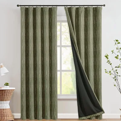 Green curtains with a subtle dotted pattern hang by a window, partially drawn to reveal a black blackout lining. A plant and a small table with a lamp are nearby.