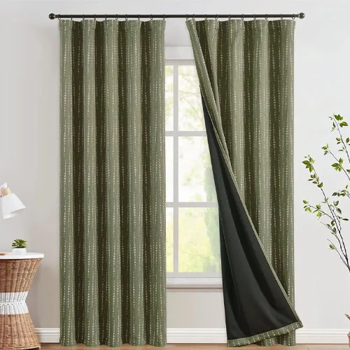 Green curtains with a subtle dotted pattern hang by a window, partially drawn to reveal a black blackout lining. A plant and a small table with a lamp are nearby.