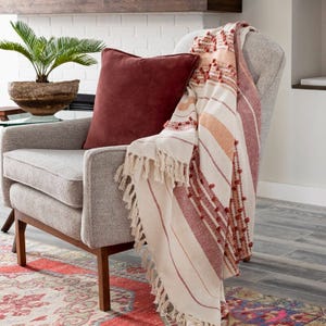 A beige upholstered armchair with wooden legs, accented by a burgundy pillow and a striped throw with fringe details.