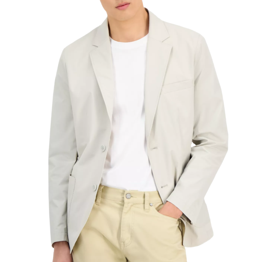 A man wearing a light gray blazer over a white T-shirt and beige pants, with one hand in his pocket.