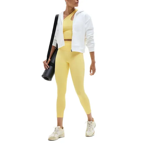 Woman wearing a yellow sports bra and leggings, white zip-up hoodie, and beige sneakers, carrying a black yoga mat.