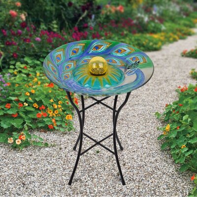 A colorful glass birdbath with peacock feather design is perched atop a simple black metal stand, positioned in a garden pathway.