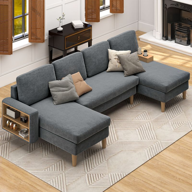 A modern gray sectional sofa with built-in storage on one armrest, adorned with various pillows. It is set on a geometric-patterned rug in a living room with wooden floors, wooden blinds, and a fireplace.