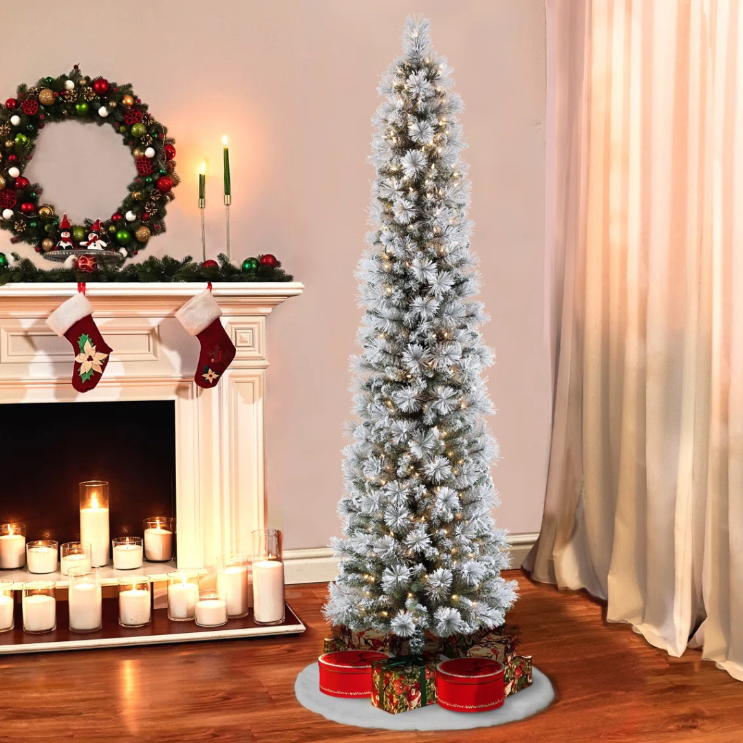 A slim, snowy artificial Christmas tree with lights is surrounded by festive gift boxes. Nearby, a decorated mantel features stockings, candles, and a wreath.