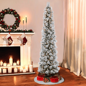 A slim, snowy artificial Christmas tree with lights is surrounded by festive gift boxes. Nearby, a decorated mantel features stockings, candles, and a wreath.
