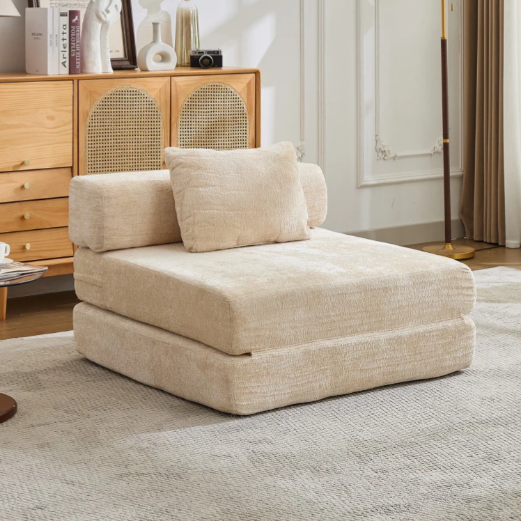 A beige, foldable futon chair with a cushion and backrest sits on a light gray rug. It's positioned in a stylish room with a wooden cabinet, decorative items, and a tall floor lamp.