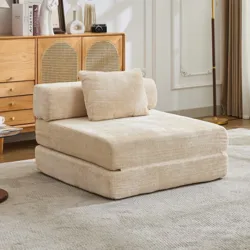 A beige, foldable futon chair with a cushion and backrest sits on a light gray rug. It's positioned in a stylish room with a wooden cabinet, decorative items, and a tall floor lamp.