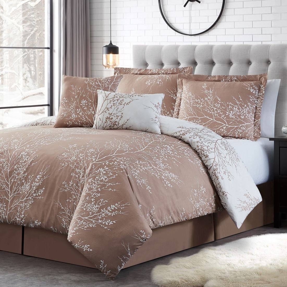 A bed with a beige and white floral-patterned comforter set, featuring pillows and a duvet, set against a tufted headboard and a brick wall with a large black clock. A pendant light hangs nearby, and a snowy landscape is visible through a window.