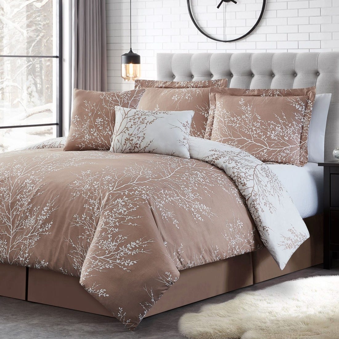 A bed with a beige and white floral-patterned comforter set, featuring pillows and a duvet, set against a tufted headboard and a brick wall with a large black clock. A pendant light hangs nearby, and a snowy landscape is visible through a window.