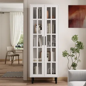White cabinet with glass-paneled doors on black legs, showcasing various books and decorative items. It's placed in a light-toned living room, near a plant, with wooden flooring and a chair in the background.