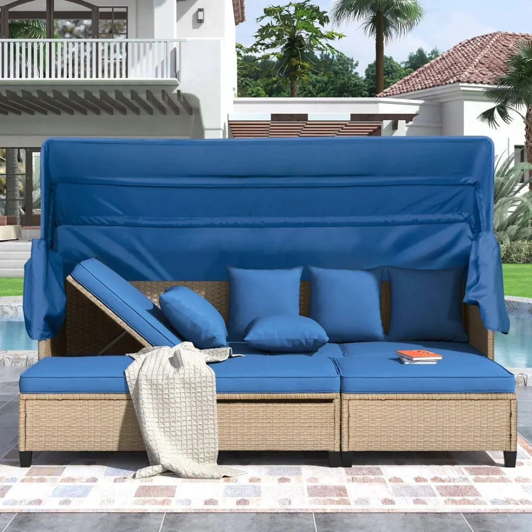 Outdoor patio daybed with an adjustable backrest, blue cushions, and canopy, set on a wicker base. Includes several matching blue throw pillows and a textured beige blanket. Pool and modern house in the background.