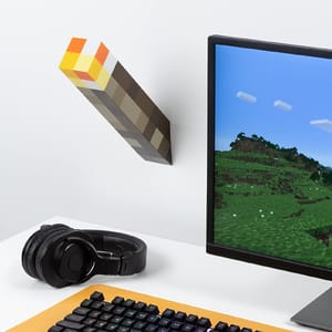 A torch-shaped wall light styled like a pixelated item next to a monitor displaying a blocky landscape, accompanied by a set of black headphones and a colorful mechanical keyboard.
