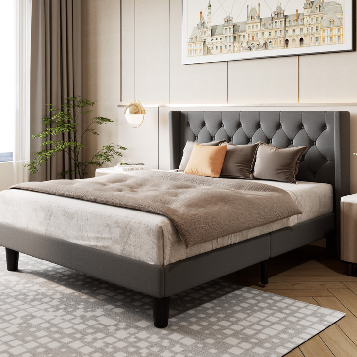 A gray upholstered platform bed with a tufted headboard. The bed is styled with neutral-colored bedding and cushions, with a modern bedside table and hanging light nearby, set in a stylishly decorated room with a plant and artwork.