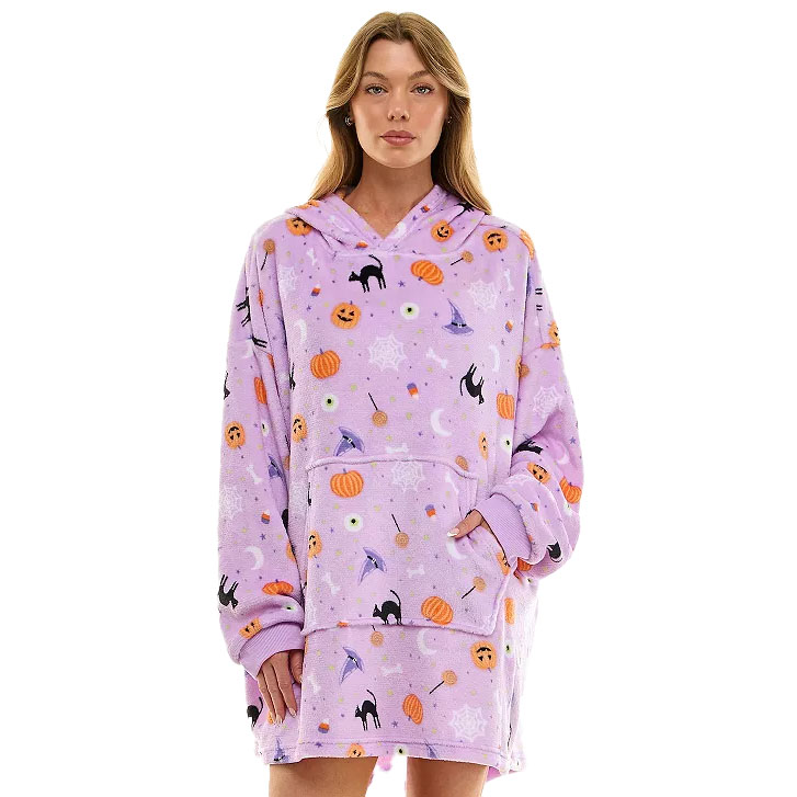 A woman wears a cozy, oversized purple hoodie blanket adorned with Halloween-themed designs, including pumpkins, bats, and cats.