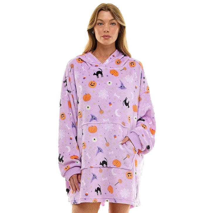 A woman wears a cozy, oversized purple hoodie blanket adorned with Halloween-themed designs, including pumpkins, bats, and cats.