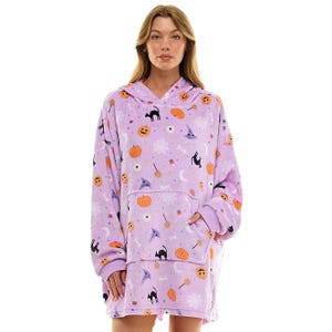 A woman wears a cozy, oversized purple hoodie blanket adorned with Halloween-themed designs, including pumpkins, bats, and cats.