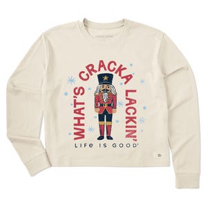Women's long sleeve shirt with a cream color featuring a nutcracker in a red and blue uniform. The text reads \“What's Cracka Lackin'\“ with \“Life is Good\“ below. Design includes light blue snowflakes.