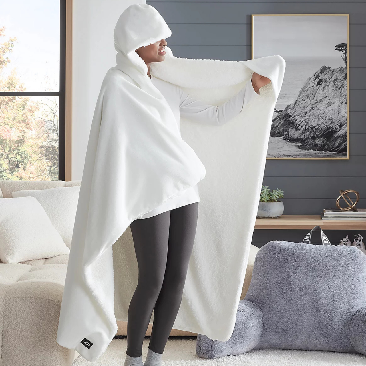A person is wearing a large, white, fleece hooded blanket with a tag labeled \“UGG.\“ The setting includes a cozy living area with plush furniture and a framed landscape artwork on the wall.