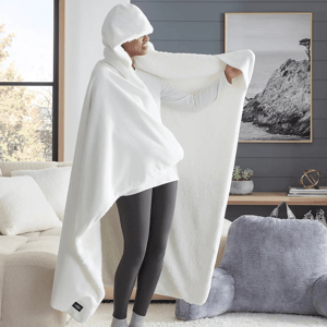 A person is wearing a large, white, fleece hooded blanket with a tag labeled \“UGG.\“ The setting includes a cozy living area with plush furniture and a framed landscape artwork on the wall.