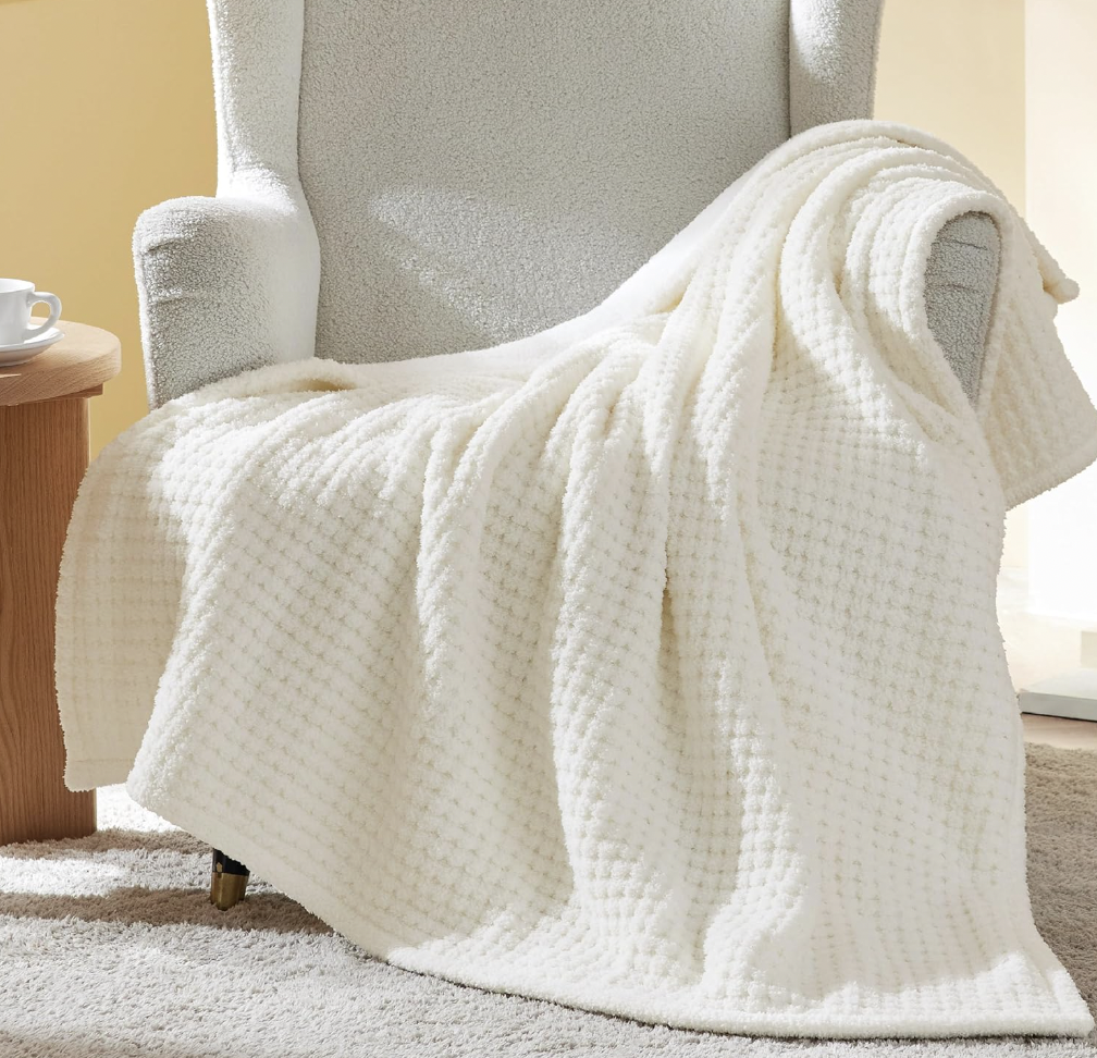 A soft, white Sherpa throw blanket draped over an armchair, featuring a textured waffle design. Nearby, a small wooden side table holds a cup and saucer, all set on a plush carpet.