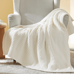 A soft, white Sherpa throw blanket draped over an armchair, featuring a textured waffle design. Nearby, a small wooden side table holds a cup and saucer, all set on a plush carpet.