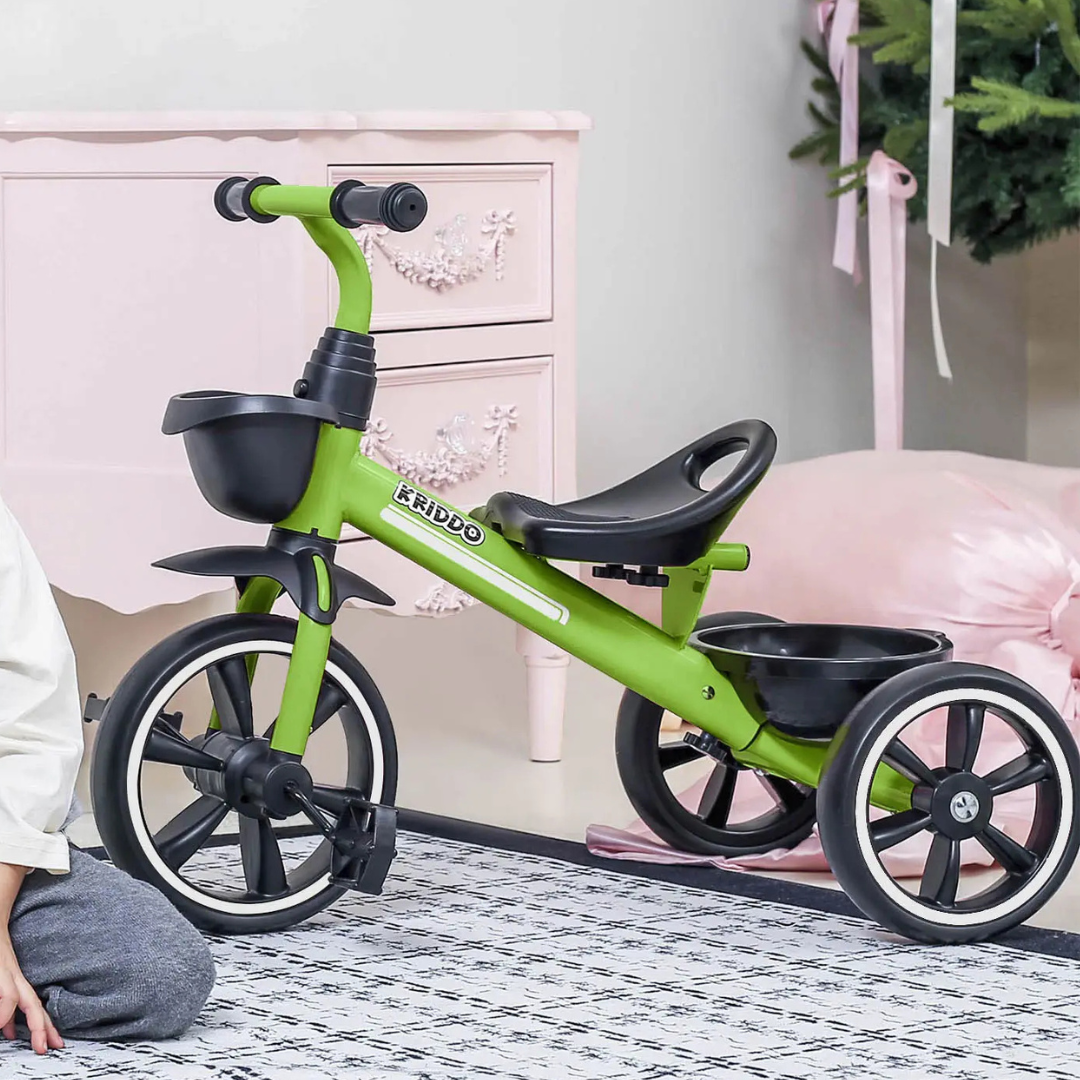 A green children's tricycle with a black seat and basket is displayed indoors, featuring sturdy wheels and handlebars.