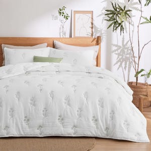 A wooden bed with a white duvet cover adorned with subtle floral embroidery, accompanied by matching pillows. A potted plant and minimalist artwork complement the serene, natural room decor.