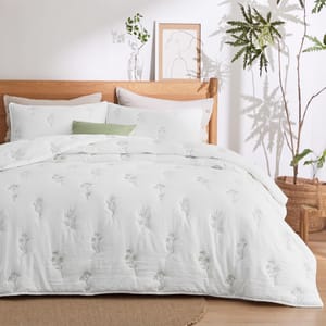 A wooden bed with a white duvet cover adorned with subtle floral embroidery, accompanied by matching pillows. A potted plant and minimalist artwork complement the serene, natural room decor.