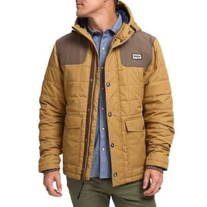 The Howler Brothers Spellbinder Parka is a quilted jacket in tan with a brown yoke and a hood. It features front pockets and a snap-button closure, paired with a blue inner shirt and olive pants.
