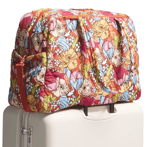 Vera Bradley Large Weekender Bag featuring a colorful floral pattern with red, blue, yellow, pink, and green tones; shown resting atop a cream-colored rolling suitcase.