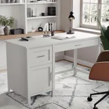 A white desk with drawers and a brown chair on castors is placed on a patterned rug. A black lamp and stationery are on the desk, and shelves with books and decor are in the background.